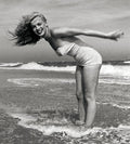 Marilyn Monroe smiling joyfully in a strapless vintage swimsuit, standing in the ocean surf with her hair blowing back.
