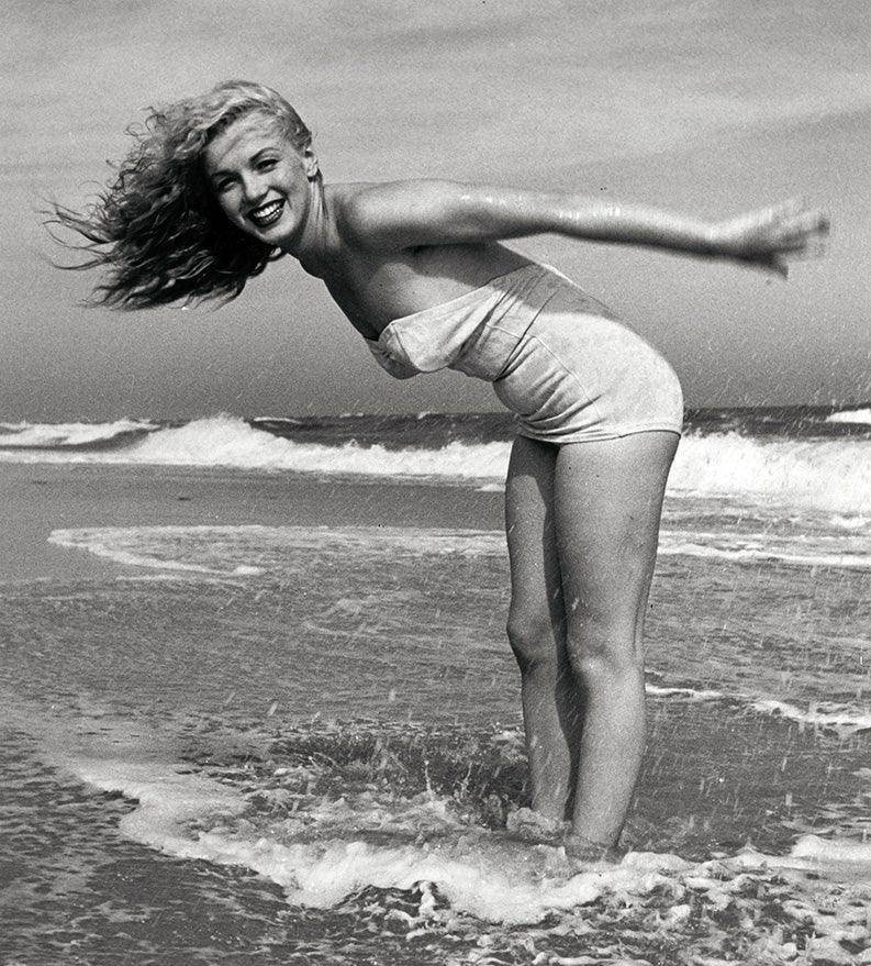 Marilyn Monroe smiling joyfully in a strapless vintage swimsuit, standing in the ocean surf with her hair blowing back.