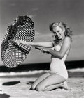Black-and-white photo of a woman in a vintage swimsuit kneeling on beach sand holding a polka dot umbrella.