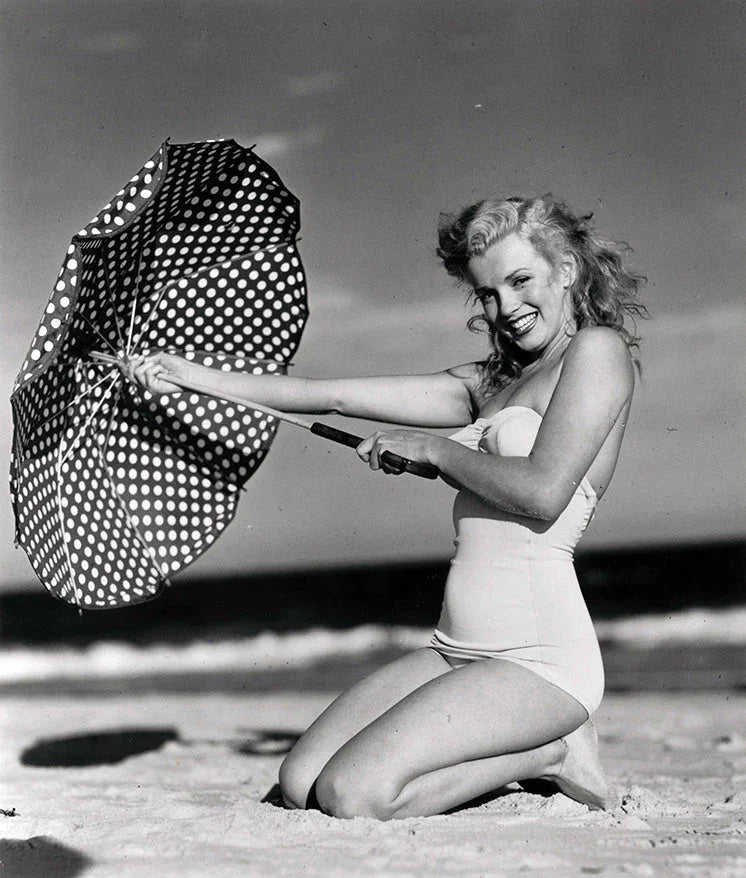 Black-and-white photo of a woman in a vintage swimsuit kneeling on beach sand holding a polka dot umbrella.