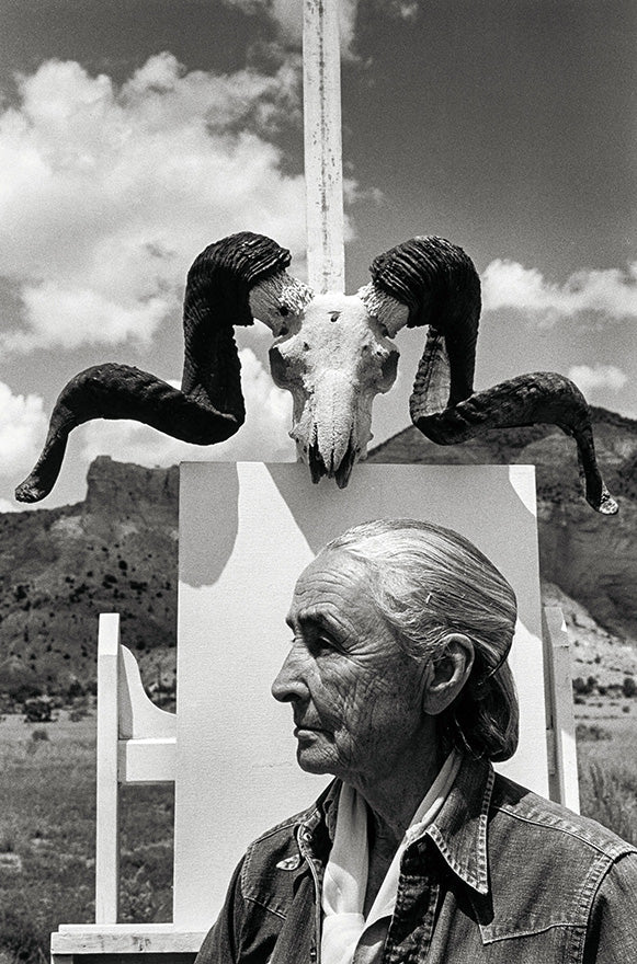 Black and white portrait of an elderly woman with silver hair in profile, seated outdoors with ram skull mounted above her on a white structure against rocky desert landscape