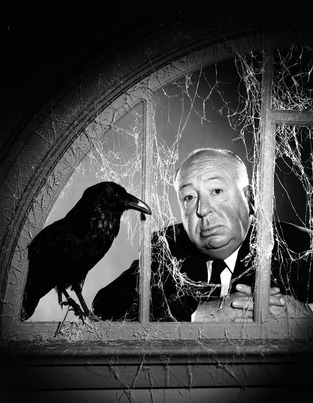 Black and white portrait of director Alfred Hitchcock leaning through a spiderweb-covered window next to a large crow, promoting his film "The Birds".