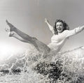 Black and white vintage photograph showing Marilyn Monroe tossed high by a splashing ocean wave, arms and legs extended in joy.