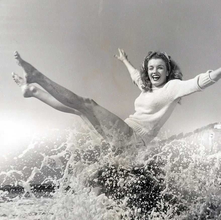 Black and white vintage photograph showing Marilyn Monroe tossed high by a splashing ocean wave, arms and legs extended in joy.
