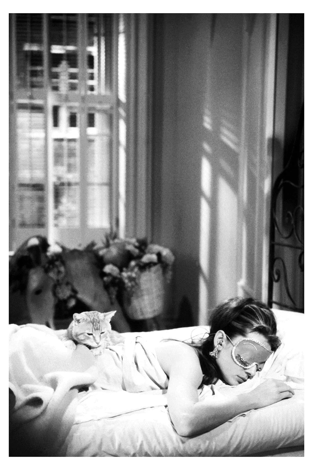 Black and white photo of woman lying on bed with eye mask and cat resting on her back