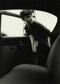 Black and white photo of Audrey Hepburn leaning gracefully out of a dark car window, wearing pearl earrings and a dark cardigan.
