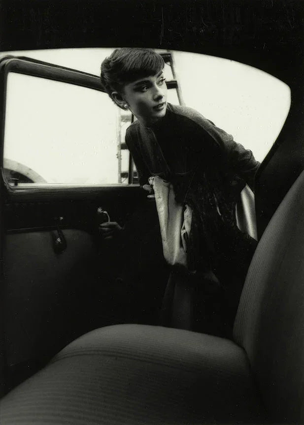 Black and white photo of Audrey Hepburn leaning gracefully out of a dark car window, wearing pearl earrings and a dark cardigan.