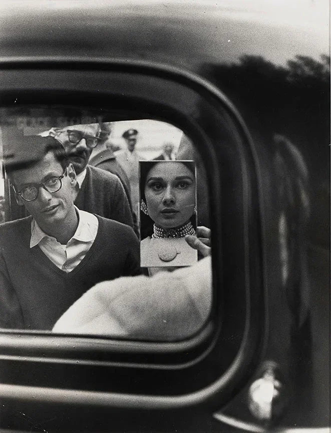 Black and white photo capturing a fan holding up a portrait of Audrey Hepburn reflected in a car window, showcasing vintage celebrity culture.