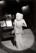 Black and white photo of Marilyn Monroe from the back, wearing the iconic sheer, glittering, backless gown while standing at a stage podium.