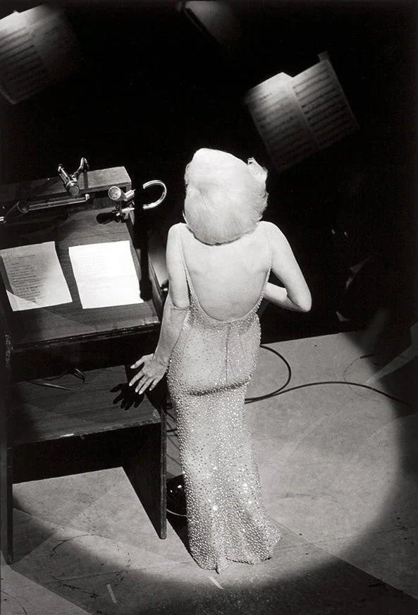 Black and white photo of Marilyn Monroe from the back, wearing the iconic sheer, glittering, backless gown while standing at a stage podium.