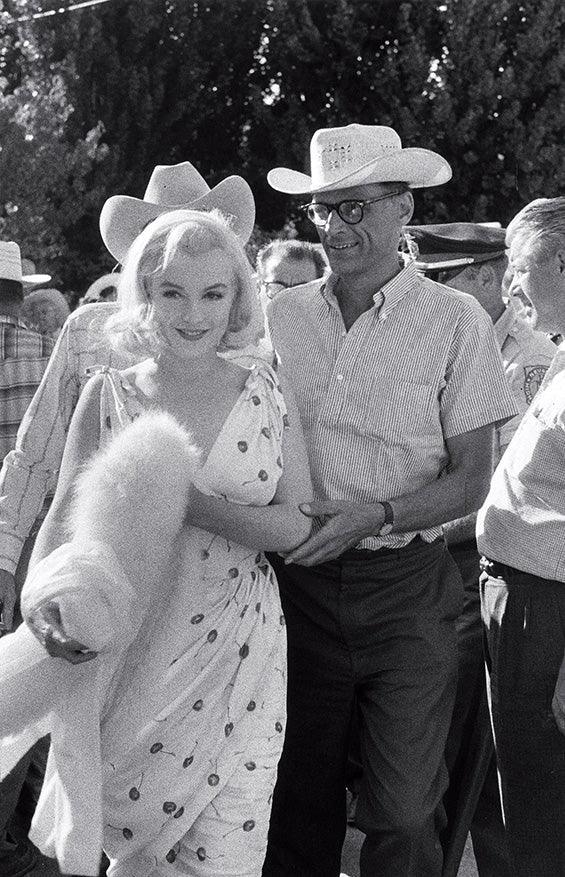 Black and white photo of a woman in cherry print dress holding a fur coat, accompanied by a man in cowboy hat and glasses outdoors