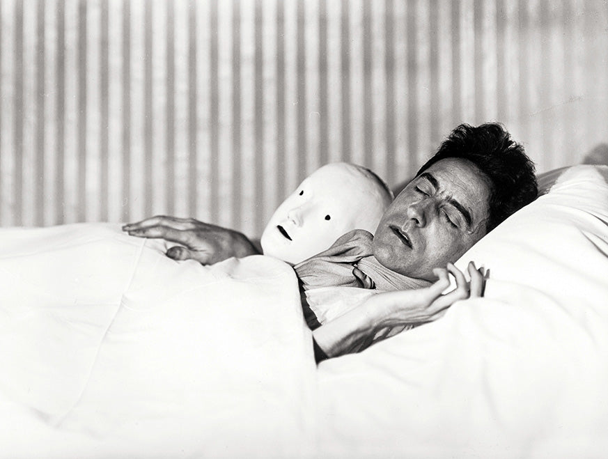 Black and white close-up photo of Jean Cocteau sleeping in bed next to a featureless white theatrical mask or mannequin head.