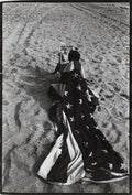 Black and white photo of a person draped in an American flag on the sand, holding up a newspaper reading "MARILYN DEAD," commemorating Marilyn Monroe's passing.