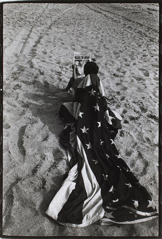 Black and white photo of a person draped in an American flag on the sand, holding up a newspaper reading "MARILYN DEAD," commemorating Marilyn Monroe's passing.