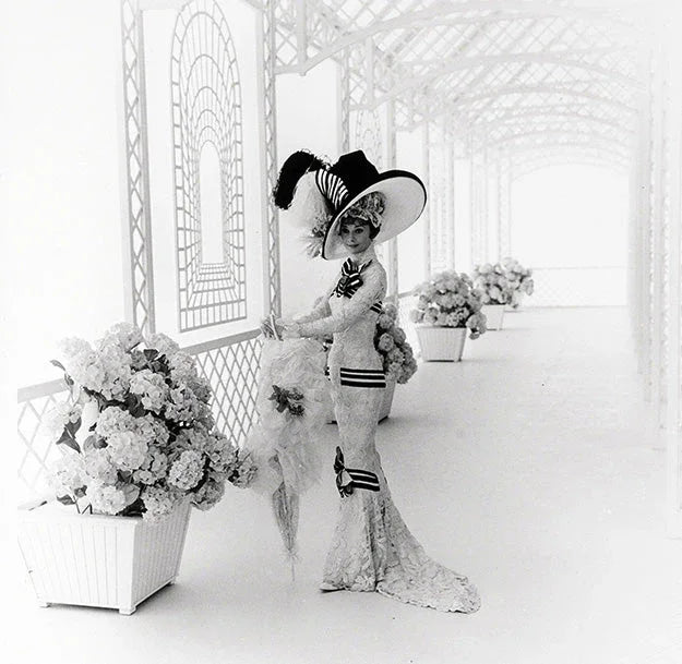 Black and white photo of Audrey Hepburn as Eliza Doolittle in a white lace gown and massive striped hat, standing among hydrangeas in a lattice corridor.