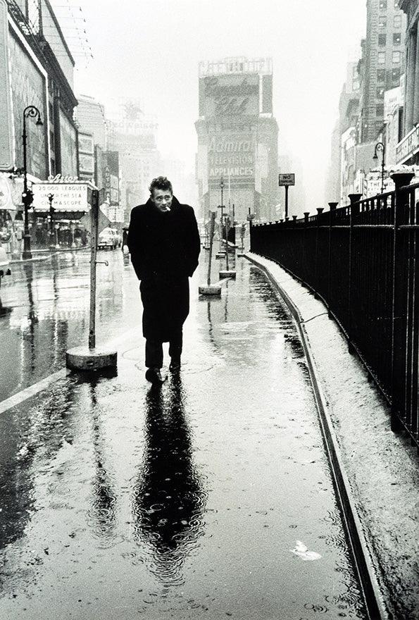 Black and white photo of James Dean walking alone on rainy Times Square sidewalk in 1955