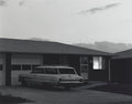 Black and white photo of a 1960s Ford station wagon parked in the driveway of a dark, mid-century ranch house at dusk.