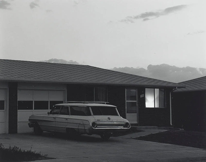 Black and white photo of a 1960s Ford station wagon parked in the driveway of a dark, mid-century ranch house at dusk.