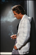 Don Johnson in profile, wearing a white blazer and lavender pants, exhaling cigarette smoke in a moody 1980s photograph.
