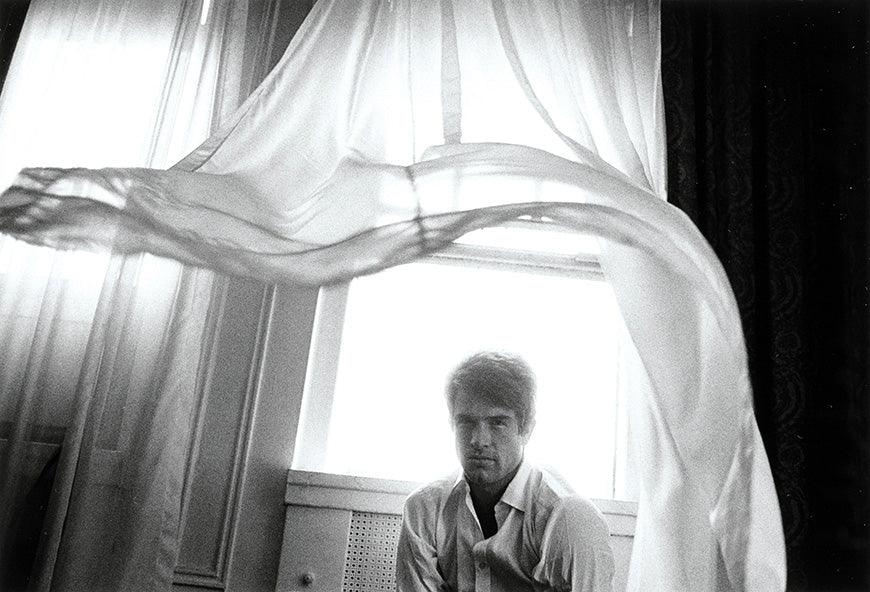 Black and white photo of a man sitting near a window with sheer curtains flowing in the wind