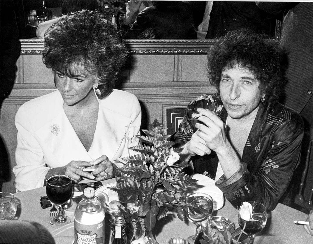 Black and white photo print showing Bob Dylan sitting at a table next to Elizabeth Taylor in 1986