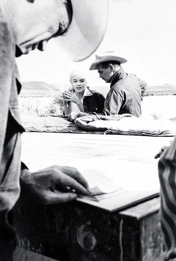 Black and white photo on the set of The Misfits featuring Marilyn Monroe and two men in cowboy hats in a desert setting