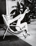 Black and white photo of smiling woman in high heels and swimsuit reclining with legs raised on chair indoors with large plants