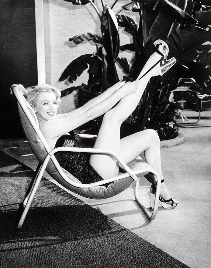 Black and white photo of smiling woman in high heels and swimsuit reclining with legs raised on chair indoors with large plants