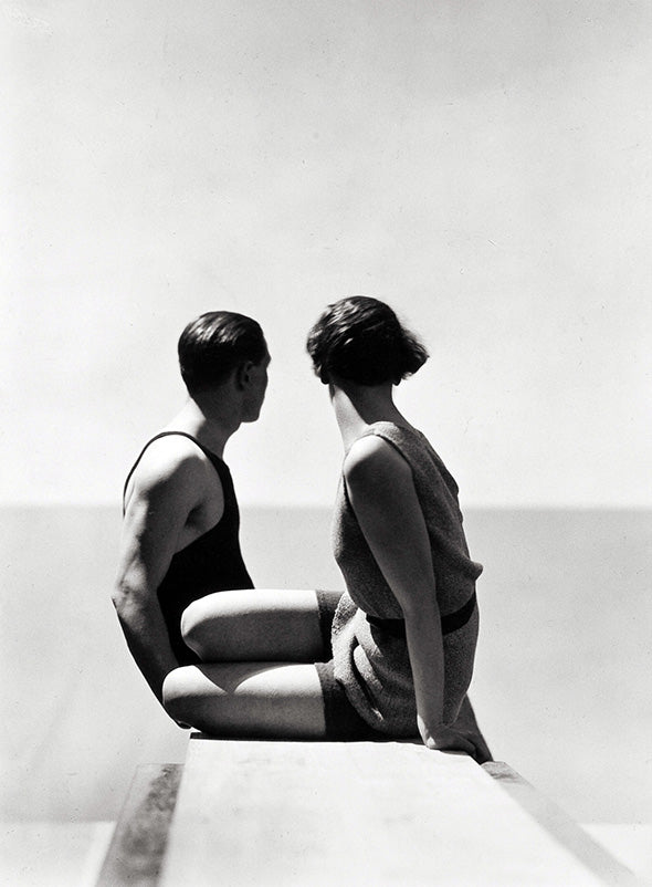High-contrast black and white photo of a 1930s couple in swimsuits sitting on a ledge looking at the ocean horizon.
