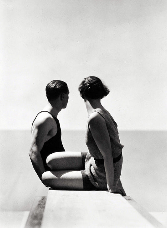 High-contrast black and white photo of a 1930s couple in swimsuits sitting on a ledge looking at the ocean horizon.