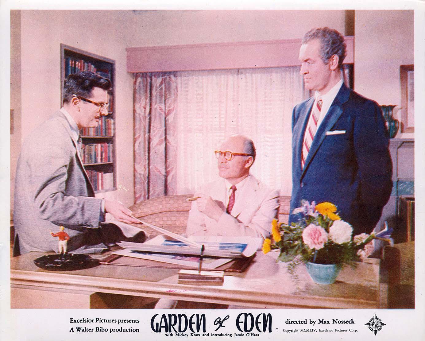 Vintage lobby card photo from the 1954 film Garden of Eden, showing three men discussing business in a formal, mid-century office environment with a subtle pink backdrop.