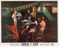 Lobby card for the 1954 movie Garden of Eden, showing a couple kissing next to a green trailer while others sit in folding chairs at night.