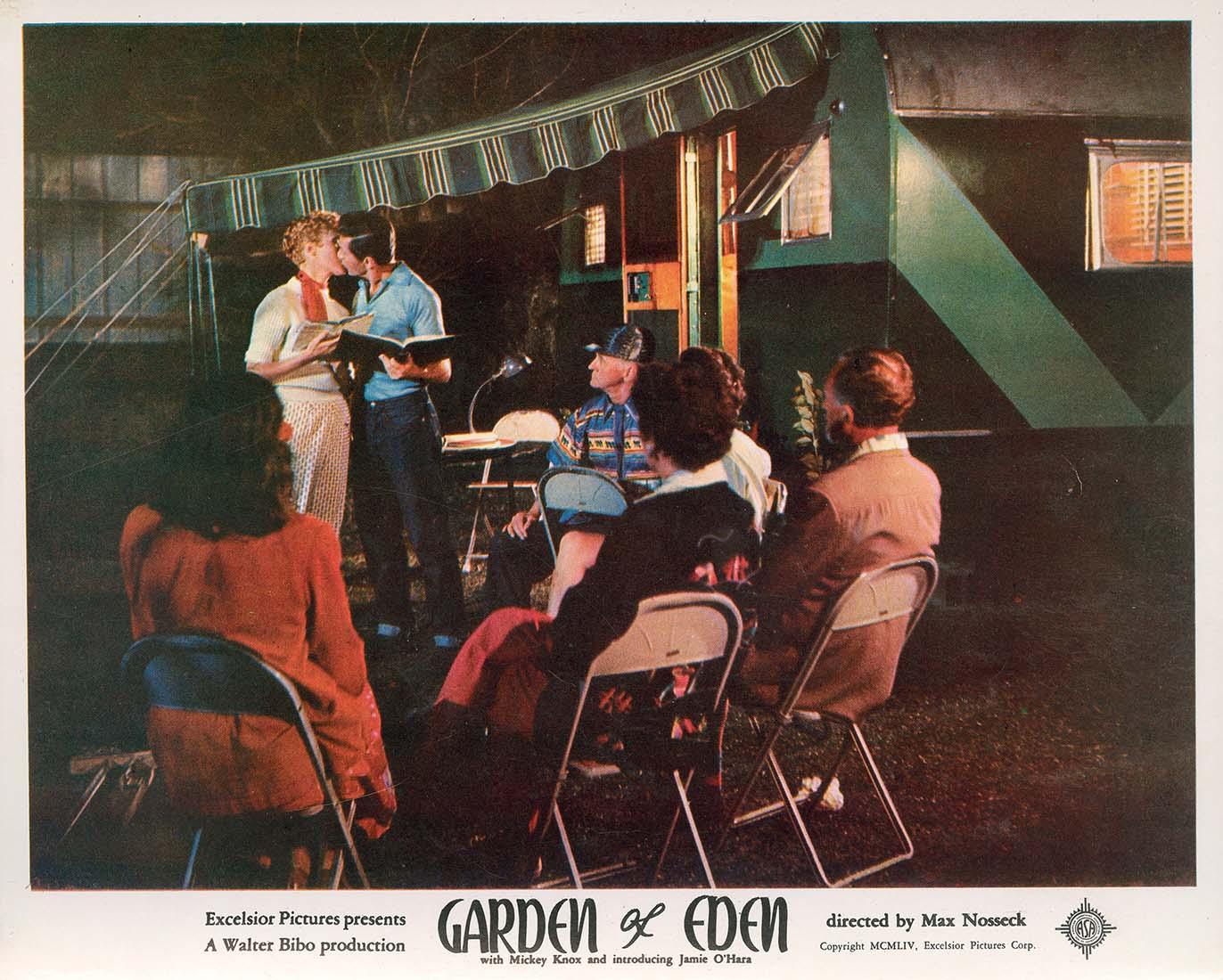 Lobby card for the 1954 movie Garden of Eden, showing a couple kissing next to a green trailer while others sit in folding chairs at night.