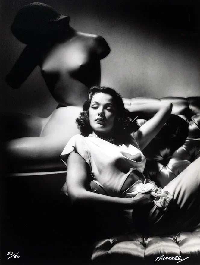 Black and white portrait of a reclining woman in elegant attire on a tufted sofa beside a large abstract sculpture