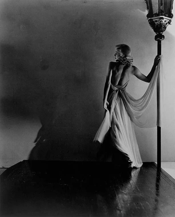 Black and white photo of a woman in a flowing, low-cut backless gown, dramatically posed holding an ornate street lamp in the style of Old Hollywood fashion photography.