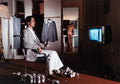 Man in pinstripe blazer sitting on chair surrounded by empty beer cans, another naked man washing in bathroom, TV showing clock