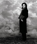 Black and white portrait of a woman in a long coat and scarf holding a book titled 'ATCHE' against a cloudy painted backdrop