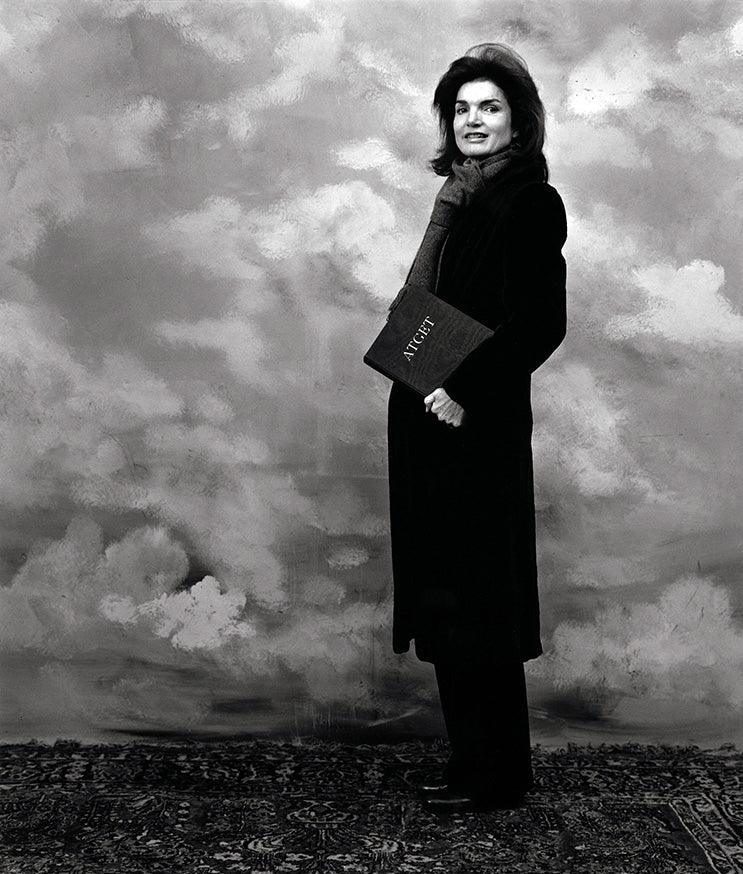 Black and white portrait of a woman in a long coat and scarf holding a book titled 'ATCHE' against a cloudy painted backdrop