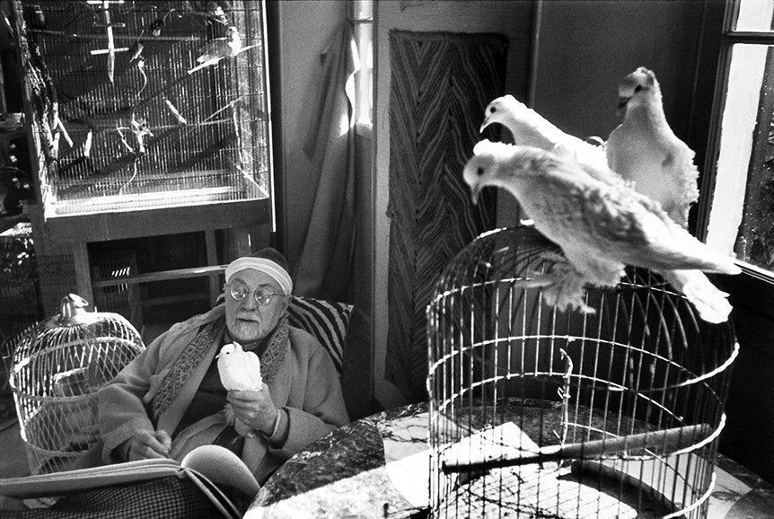Black and white photo of an elderly man with glasses and headband holding a white bird indoors surrounded by birdcages