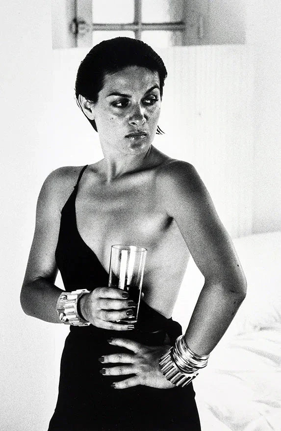 Black and white photo of a woman in a one-strap dress holding a glass, wearing multiple bracelets, in a bright room