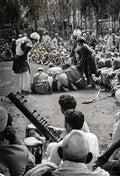Black and white photo of a large outdoor gathering in Kashmir with men and boys watching a traditional street performance and sitar music