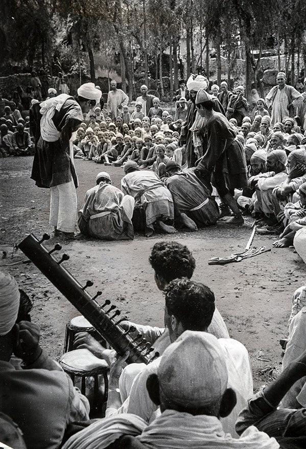 Black and white photo of a large outdoor gathering in Kashmir with men and boys watching a traditional street performance and sitar music