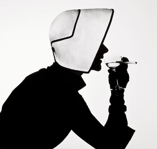 Striking black and white fashion photo of a woman in silhouette wearing a dramatic, architectural bonnet while holding a cigarette and a martini glass.