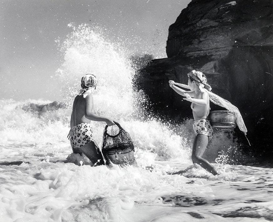 Black and white photo of two women in patterned swimwear and headscarves fishing with nets in ocean waves near rocky shore