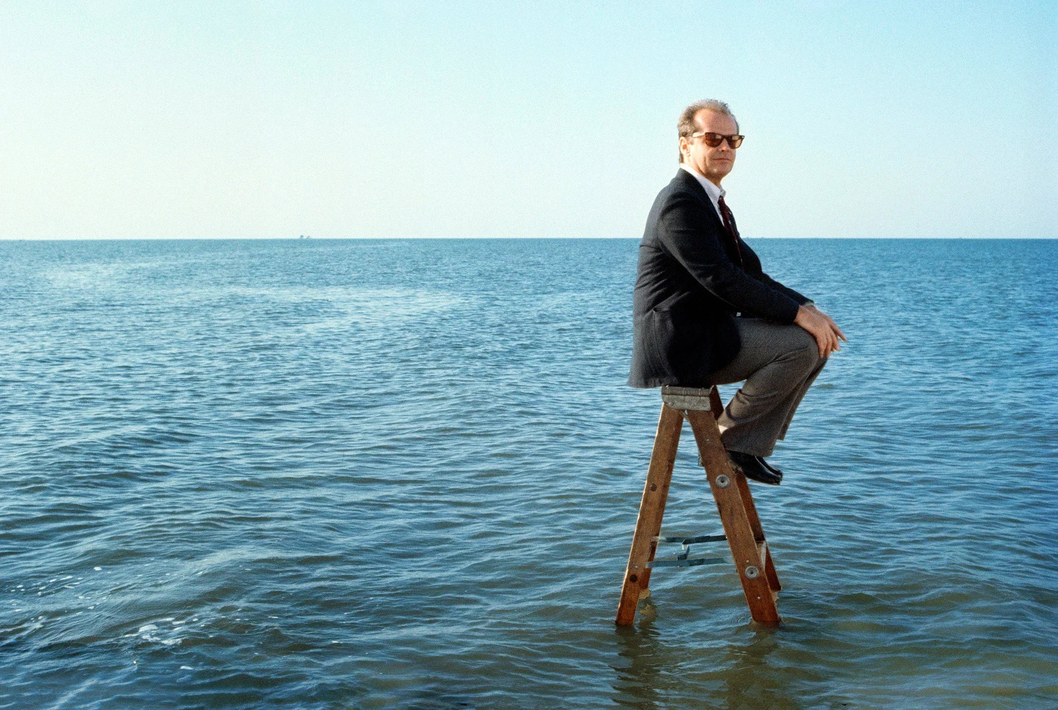 Jack Nicholson, wearing a suit and sunglasses, sitting contemplatively on a wooden ladder placed in calm, blue ocean water. Surreal actor fine art print.