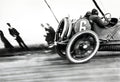 Historic black and white photo capturing a vintage open-wheel race car (No. 6) speeding past blurred spectators, emphasizing motion and early 20th-century motor racing.