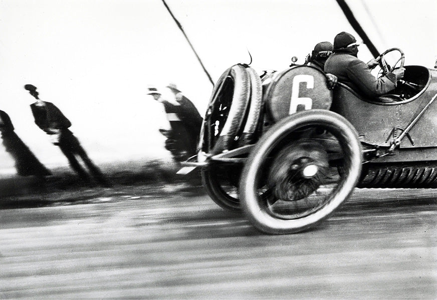 Historic black and white photo capturing a vintage open-wheel race car (No. 6) speeding past blurred spectators, emphasizing motion and early 20th-century motor racing.