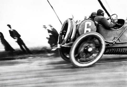 Historic black and white photo capturing a vintage open-wheel race car (No. 6) speeding past blurred spectators, emphasizing motion and early 20th-century motor racing.