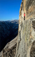 Climber standing on narrow ledge on Yosemite Half Dome cliff with vast mountain valley and blue sky