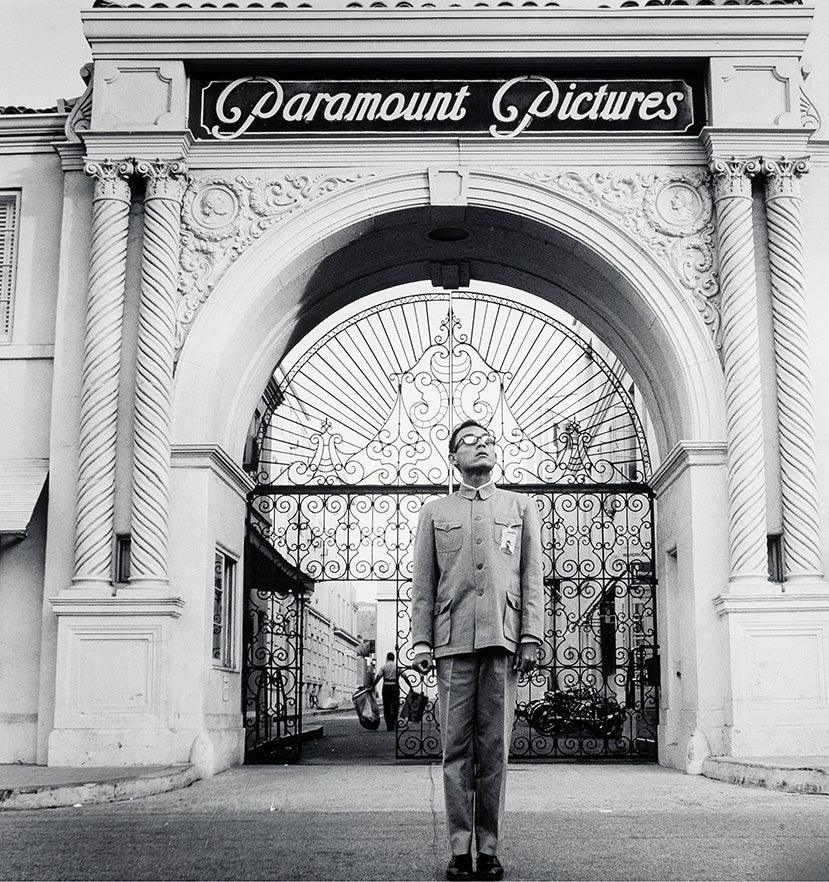 Black and white photo of a man in uniform standing in front of ornate Paramount Pictures entrance gate in Los Angeles